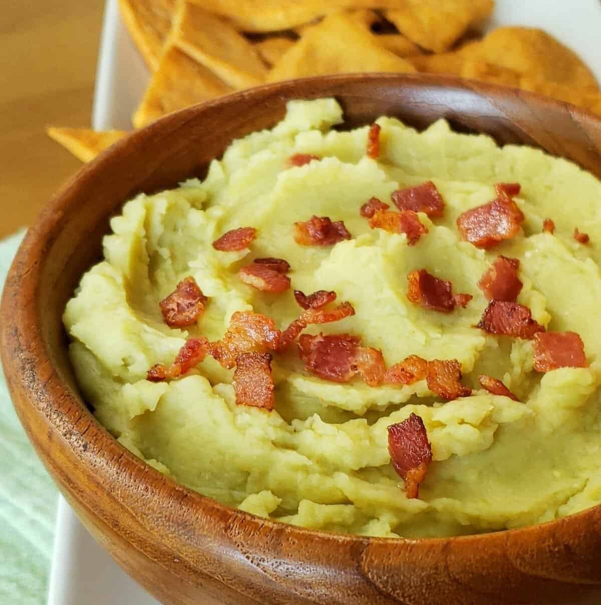 Wooden bowl of lima bean dip with bacon sprinkled on top and pita chips in background.