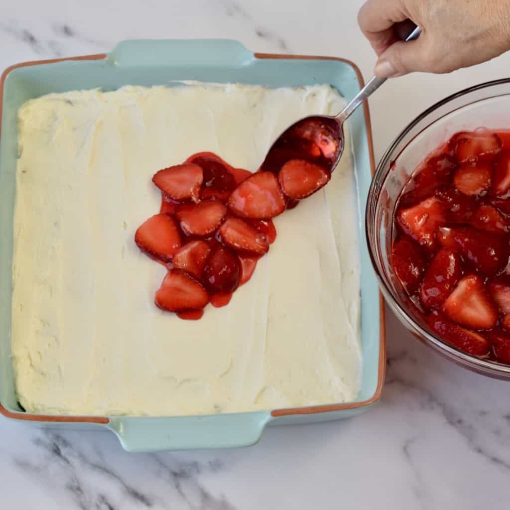 Hand spooning strawberry mixture onto cream cheese mixture in a blue 8x8 pan.