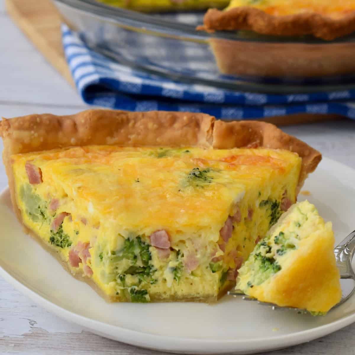 Slice of ham broccoli quiche on white plate with glass pie plate in background on blue cloth.