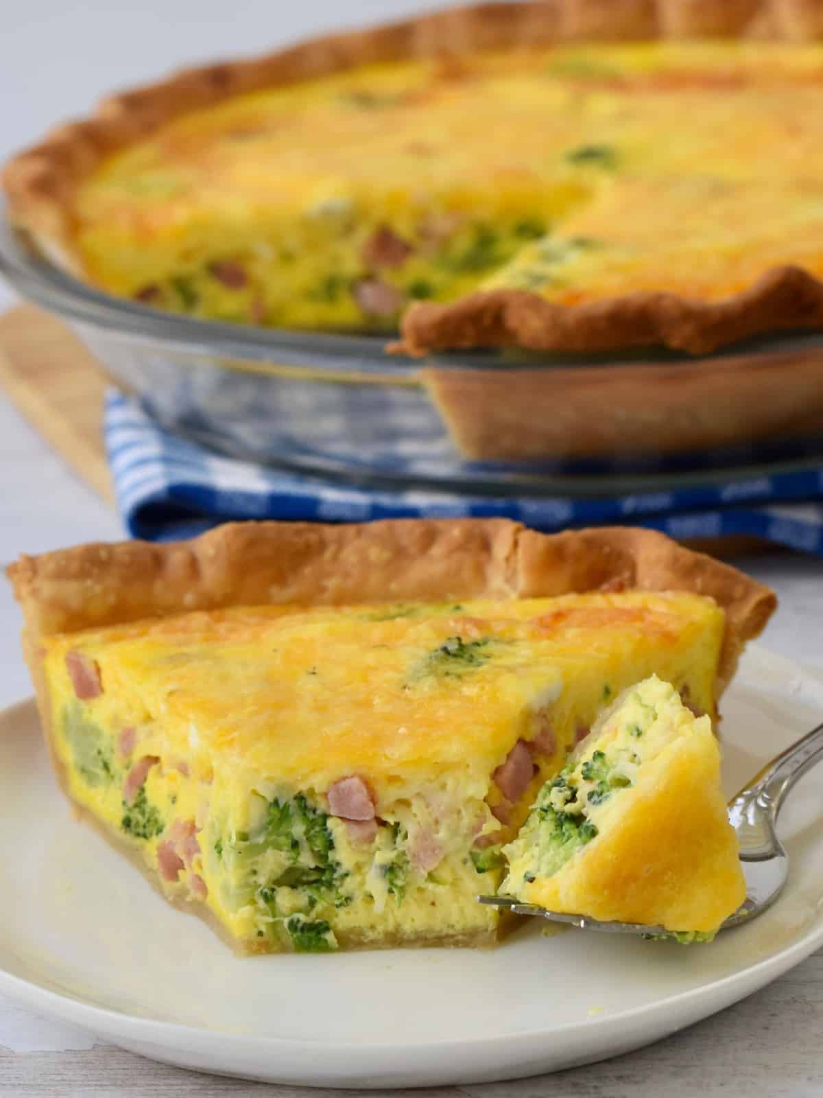 Ham Broccoli Quiche bite out on fork with quiche in background on blue checked napkin.