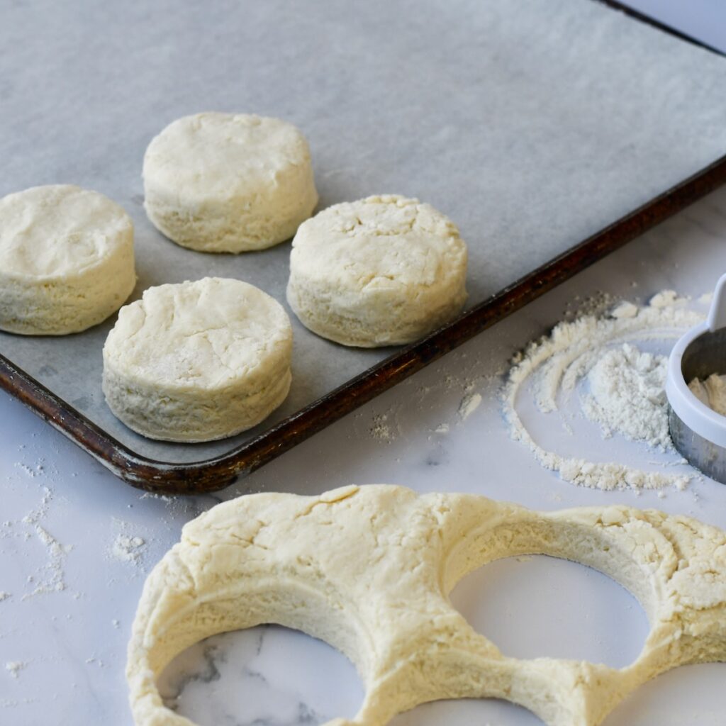 Uncooked homemade biscuits on a baking sheet and on marble surface.