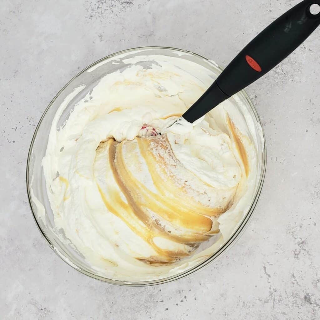 Black spatula folding peanut butter into whipped cream in glass bowl.