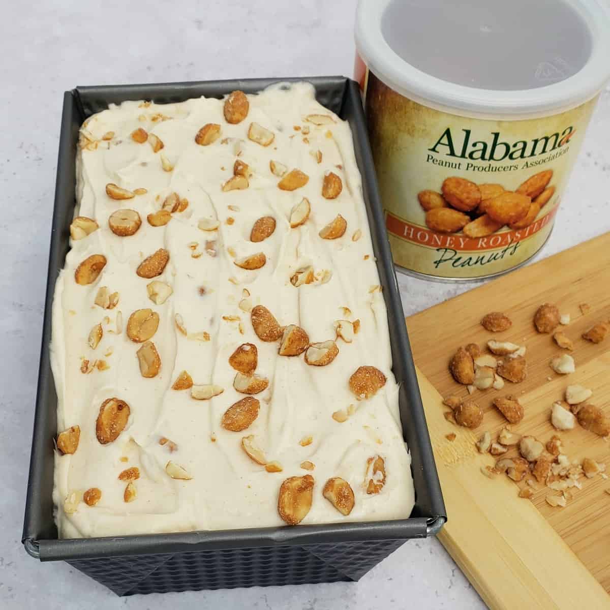 Loaf pan full of peanut butter ice cream topped with honey roasted peanuts with can of peanuts next to it.