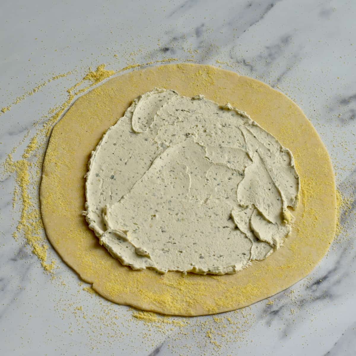 Herbed cream cheese spread over most of flat pie crust on marble surface.