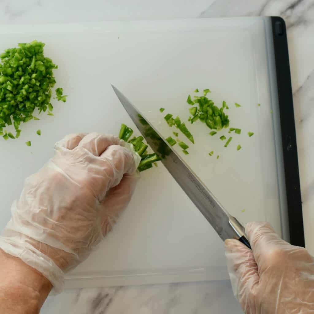 Hands mincing jalapeno peppers with chefs knife wearing latex gloves
