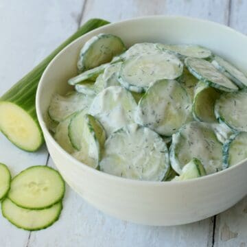 White bowl of creamy cucumber salad with half sliced cucumber next to it.