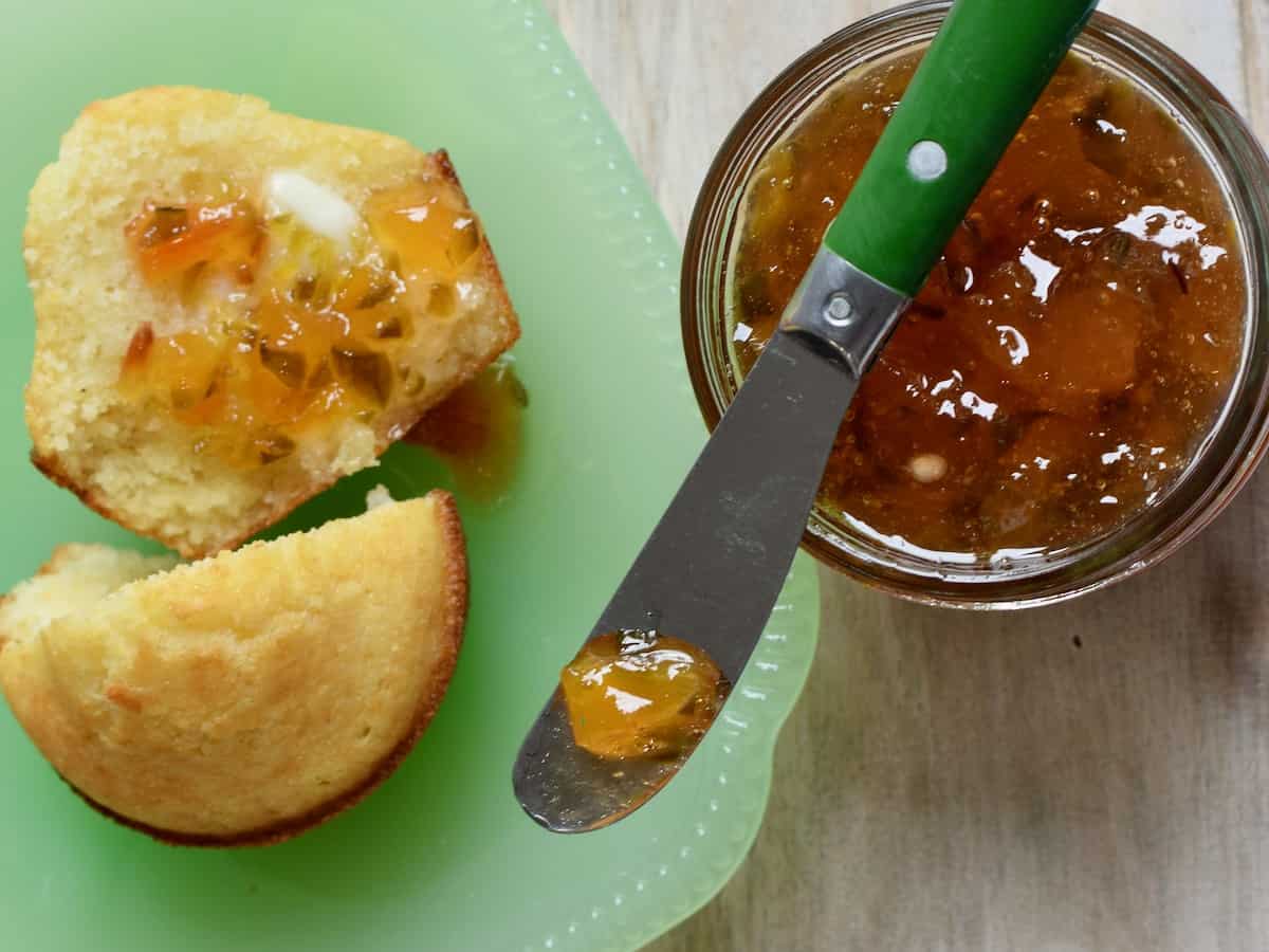 Cornbread muffin with peach pepper jelly on green plate and jar of jelly with spreader.