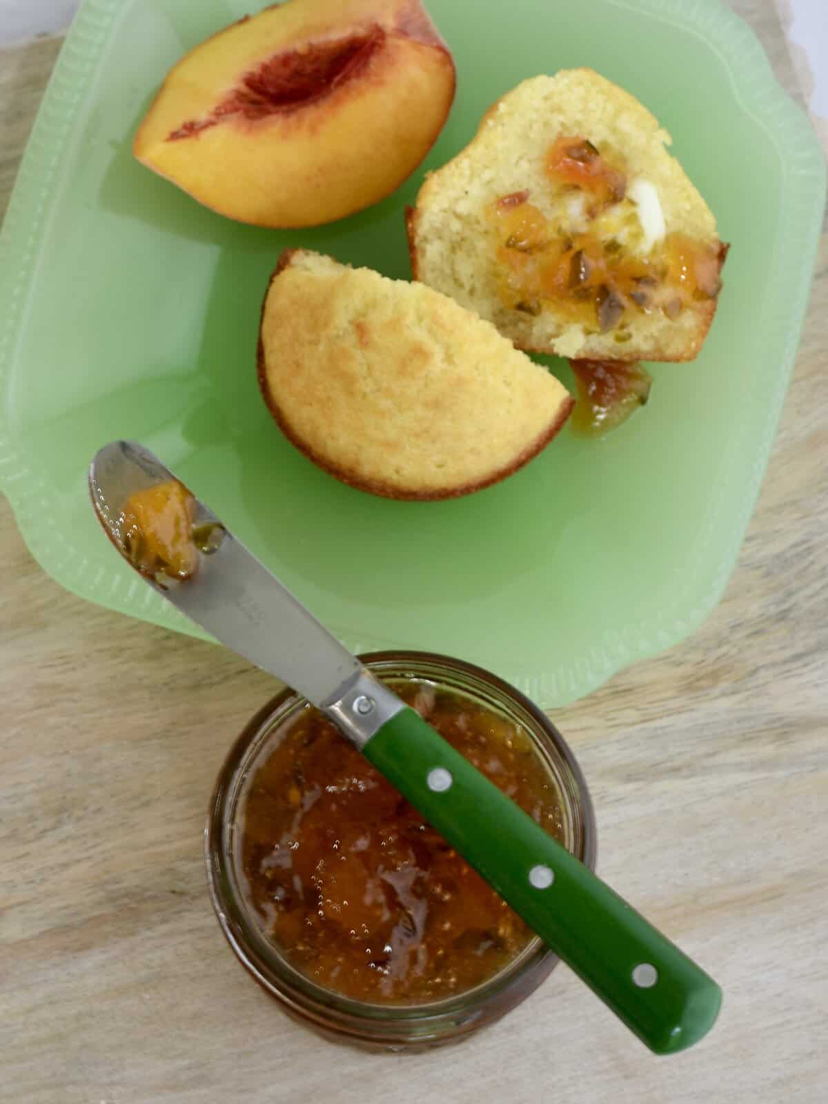 Cornbread muffin with peach pepper jelly on one half; jar of jelly and spreader at the bottom.