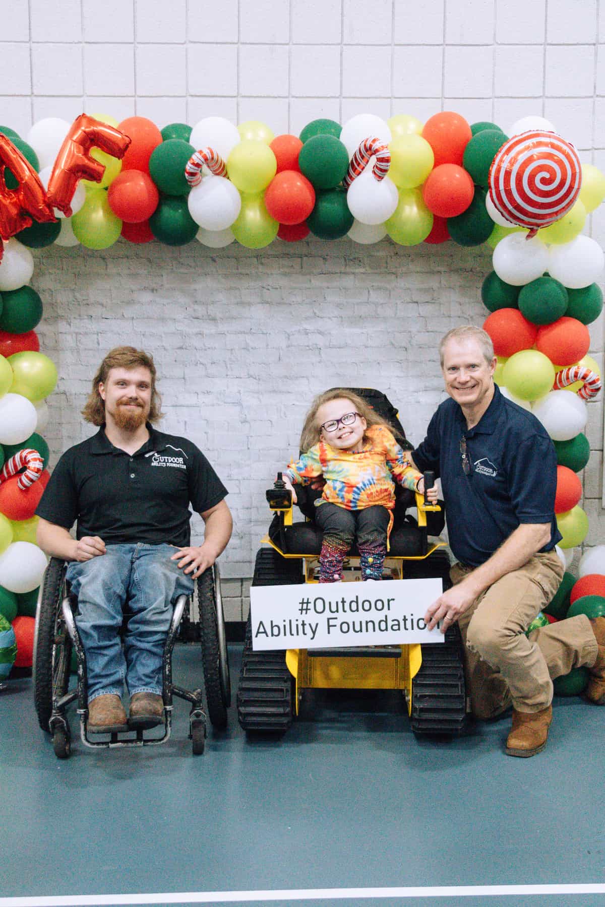 Man with beard in wheelchair next to little girl in wheelchair with man kneeling.