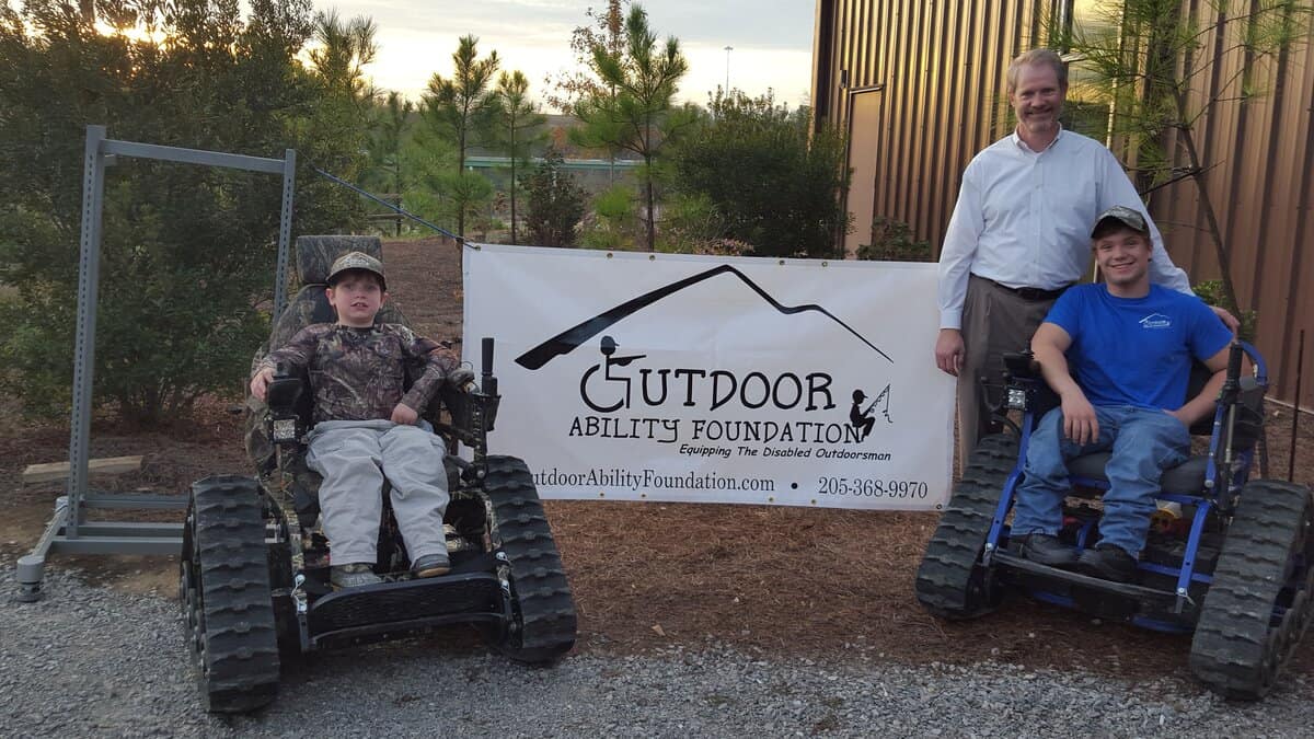 Two boys in a tank style wheelchair on either side of a banner with man standing behind one.