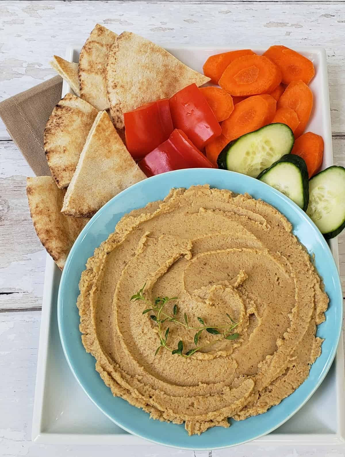 Boiled peanut hummus in blue bowl with vegetables and pita bread around it.