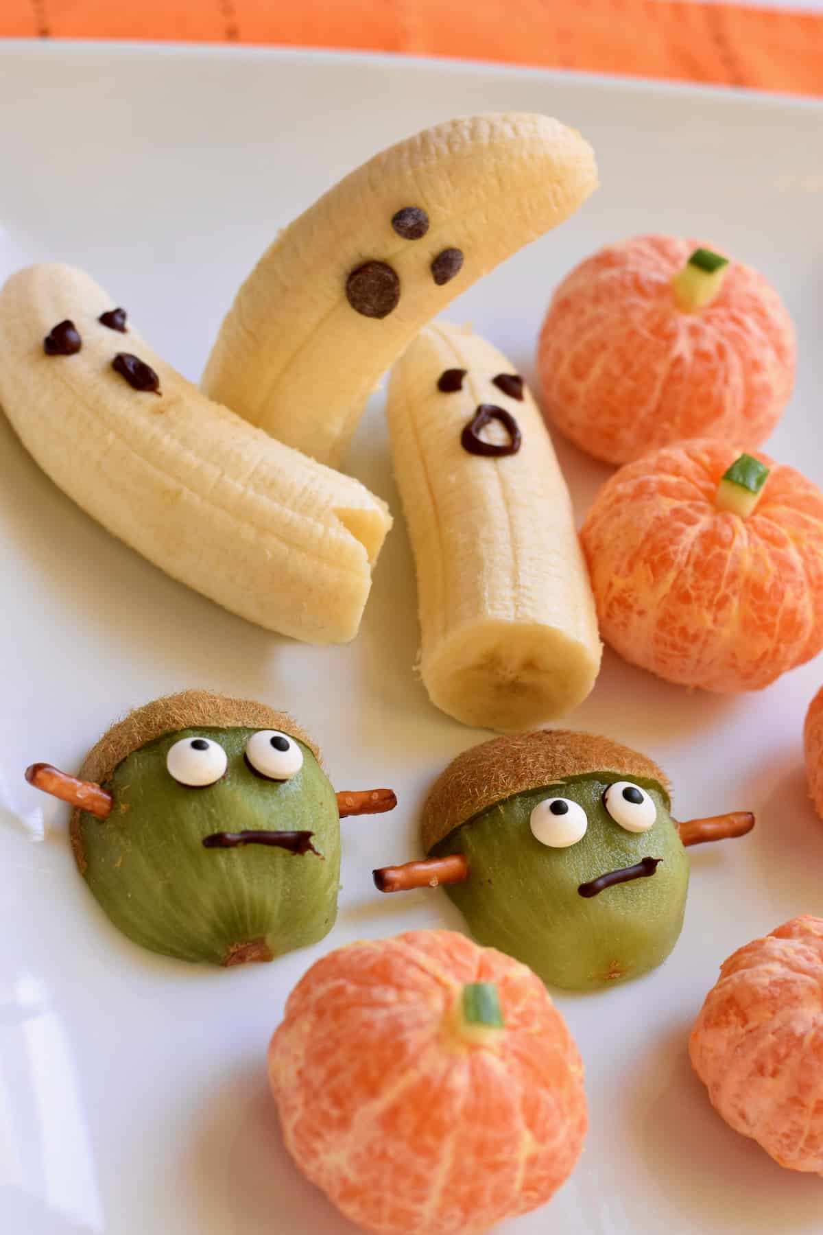 White tray of Halloween fruit snacks: banana ghosts, Frankenstein kiwi, pumpkin oranges.