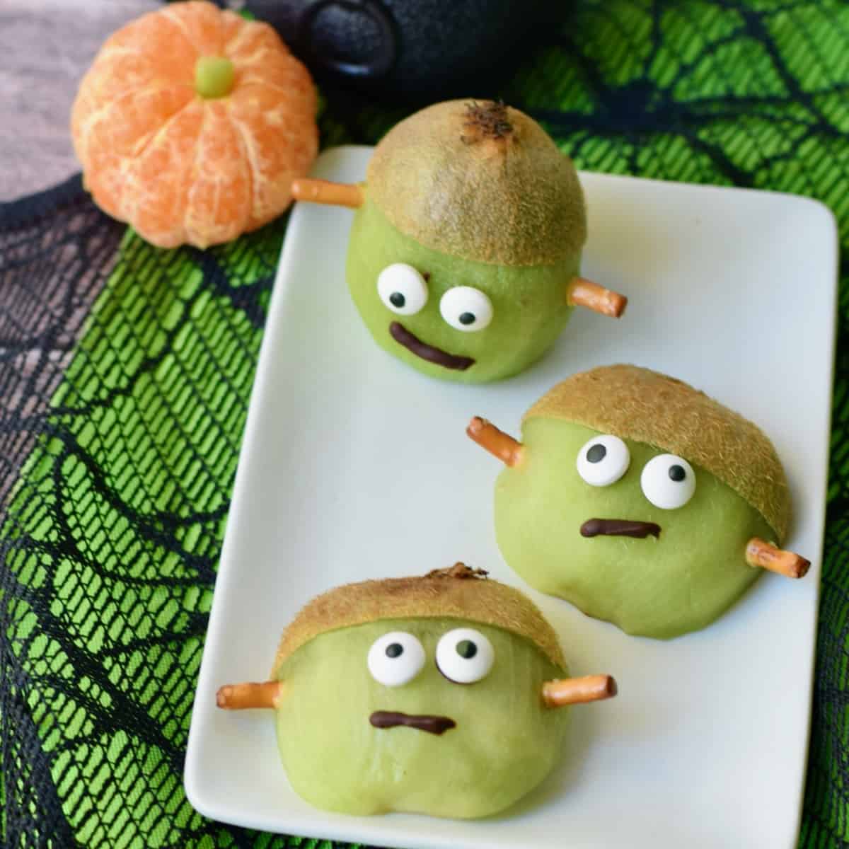 Three Frankenstein kiwi treats with chocolate mouths and candy eyes on rectangle plate..