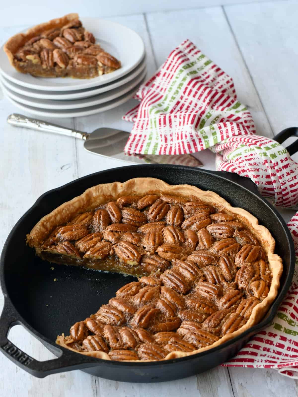 Pecan pie in cast iron skillet with slice out. Slice on top of white stack of plates.