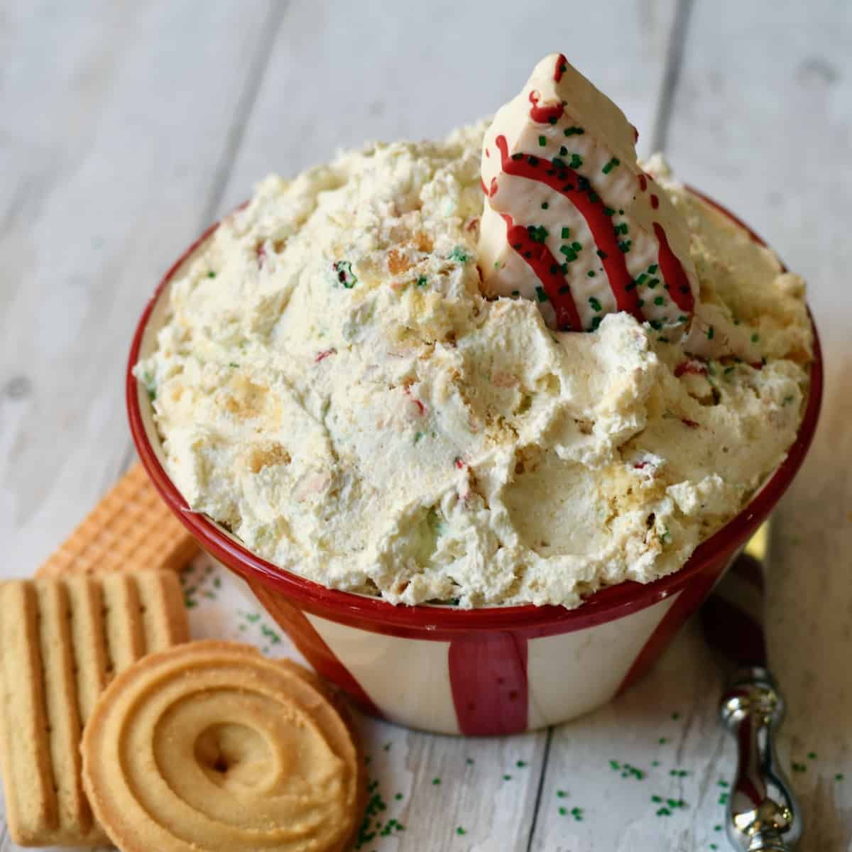 Little Debbie Christmas tree cake sticking out of red striped bowl of dip with cookies on white wood surface and metal spreader..