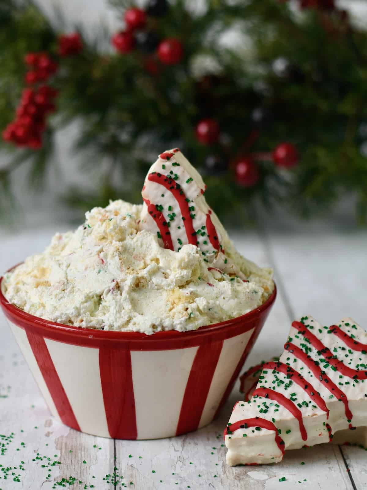 Little Debbie Christmas tree cake sticking out of red striped bowl of dip with tree cake on white wood surface and green Christmas tree in background..