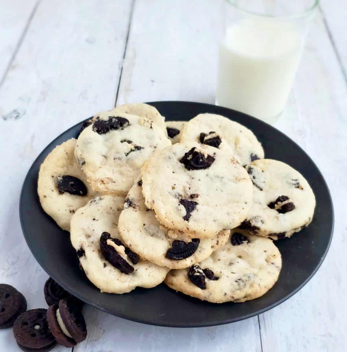 Black plate of Cookies and Cream Shortbread cookies and bite size chocolate sandwich cookies on the side with glass of milk.