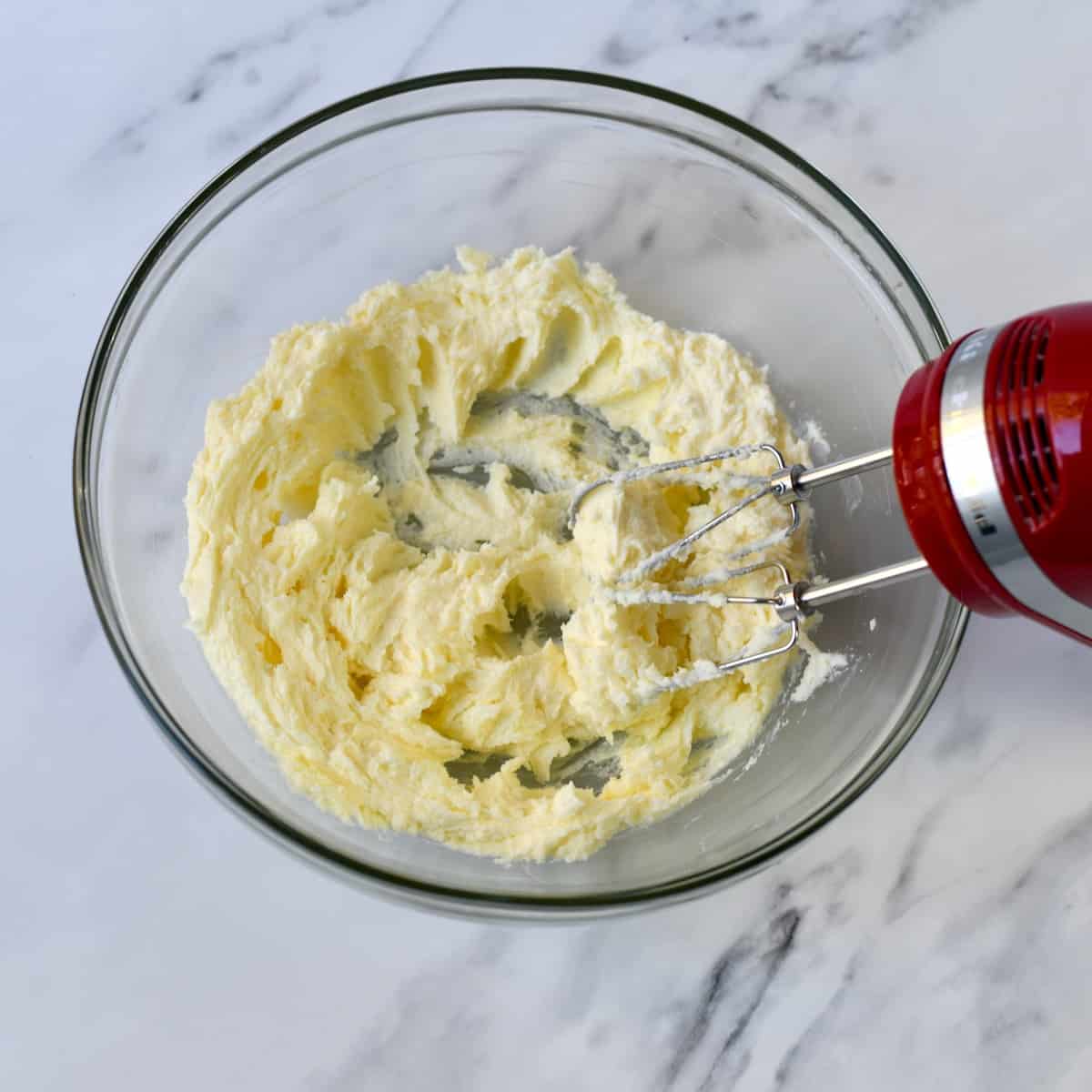Red hand mixer beating butter and sugar in a glass bowl.