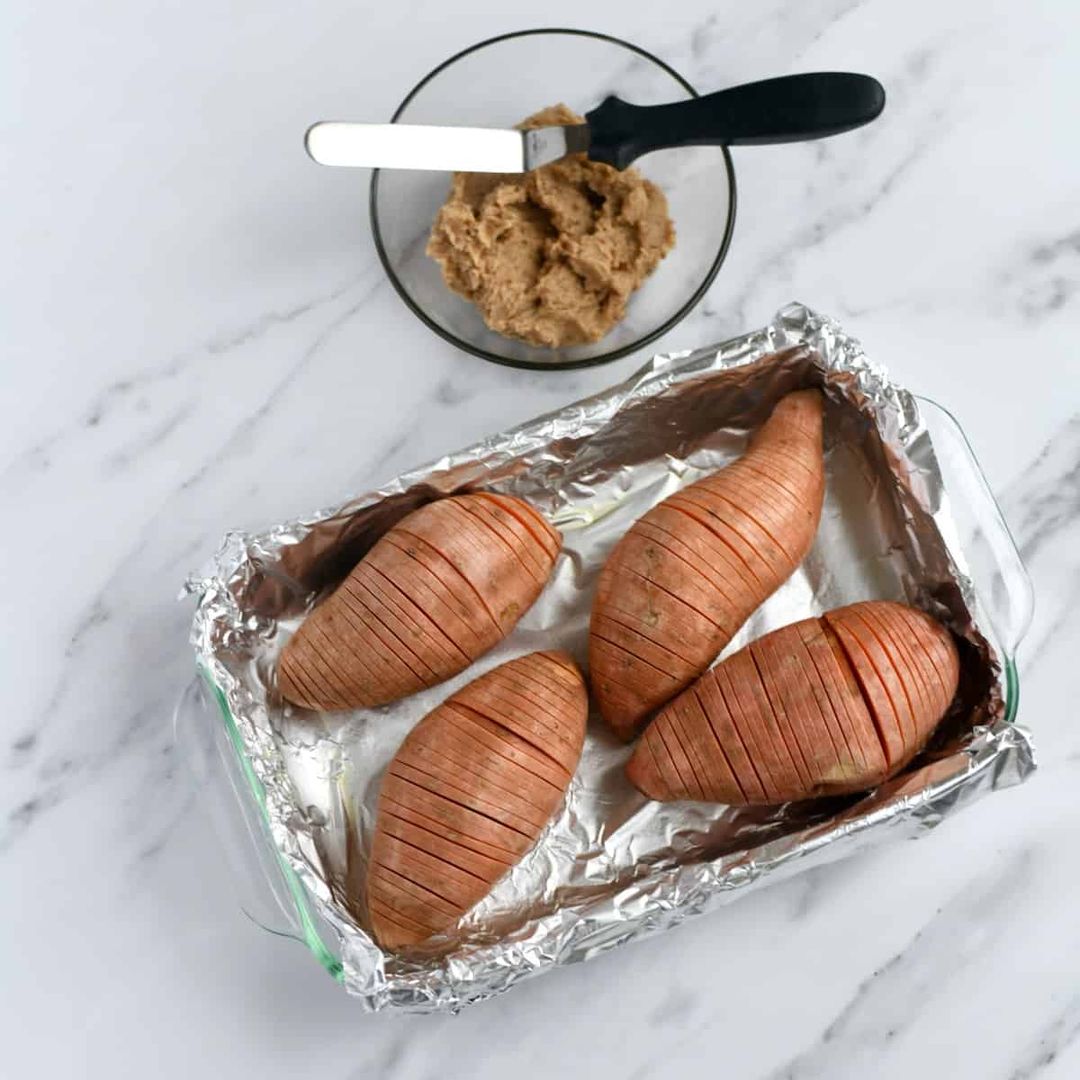 Aluminum foil lined dish with hasselback sweet potatoes in it. Glass bowl of maple pecan butter.