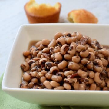 Square white bowl of black eyed peas with torn corn bread in background.