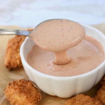 Metal spoon with comeback sauce dripping into white bowl with chicken nuggets on surface.