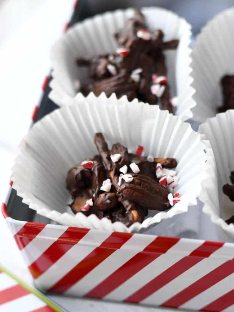 Dark chocolate pecan clusters with pretzel sticks and caramel bits in a red white striped gift tin with crushed peppermint sprinkled on top.
