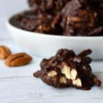Dark chocolate cluster of pecans pretzels with bite out and bowl of clusters behind it.
