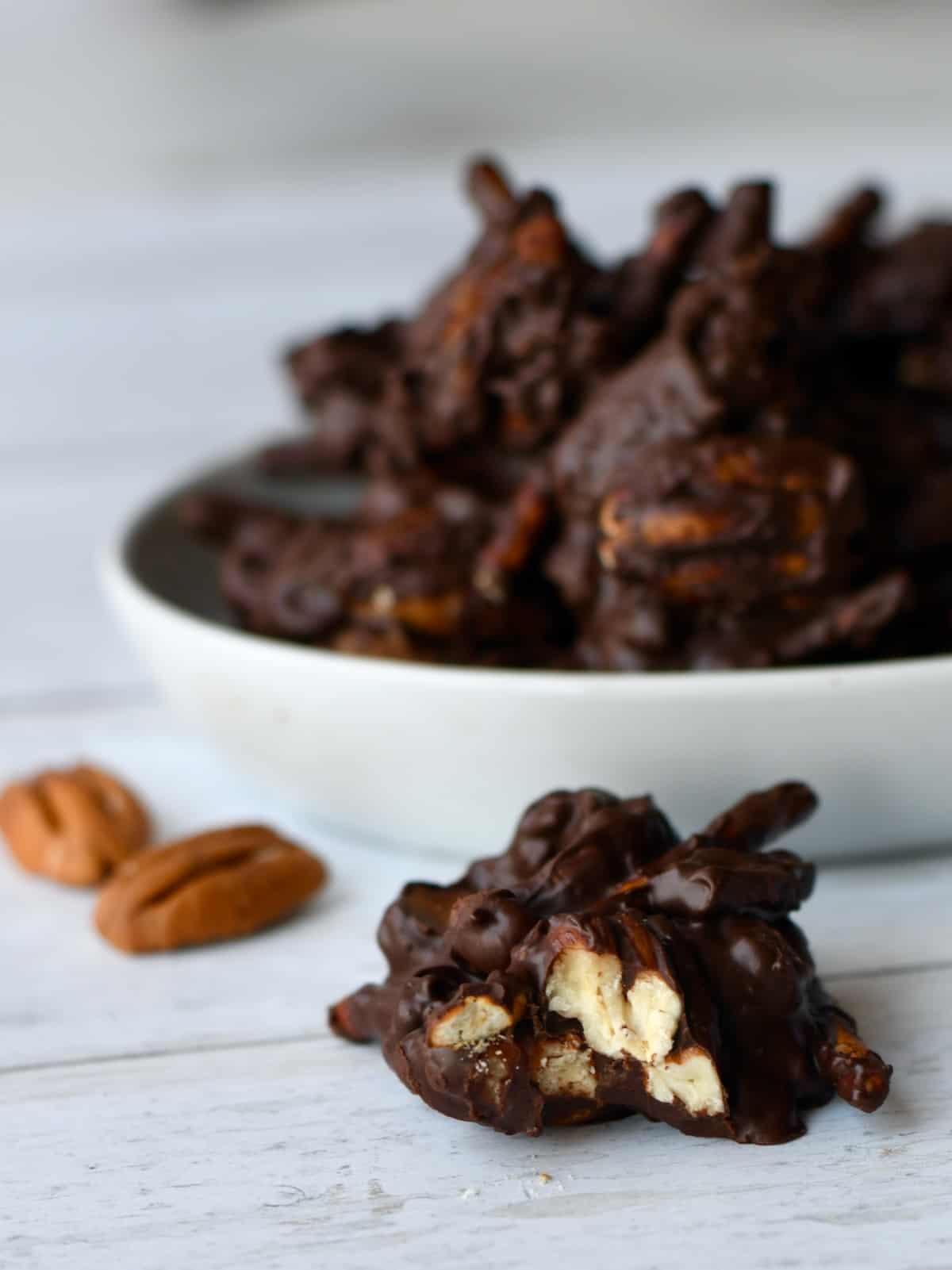 Dark chocolate cluster of pecans pretzels with bite out and bowl of clusters behind it.