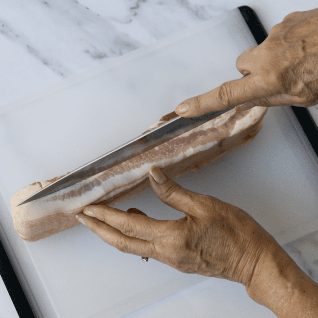 Thick-cut bacon joint on a chopping board being cut lengthwise into 2 pieces with a sharp kitchen knife