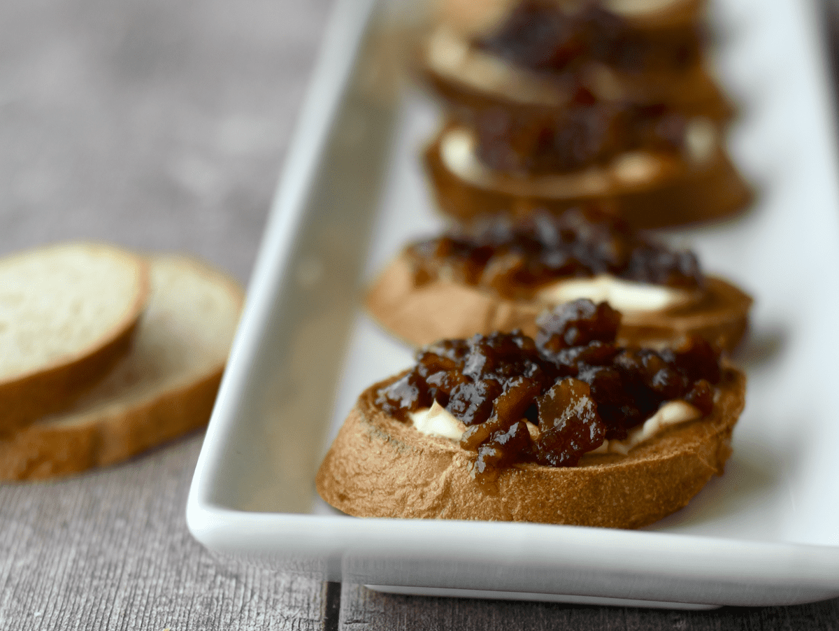 Close up image of crostini topped with cream cheese and instant pot bacon jam on a white platter