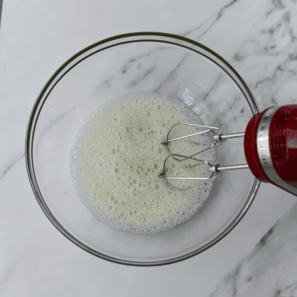 Hand mixer in glass bowl of frothy egg whites.
