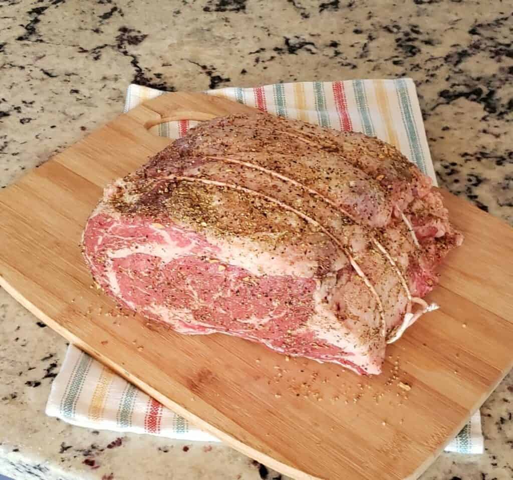 Uncooked beef ribeye roast on wooden cutting board seasoned on top.
