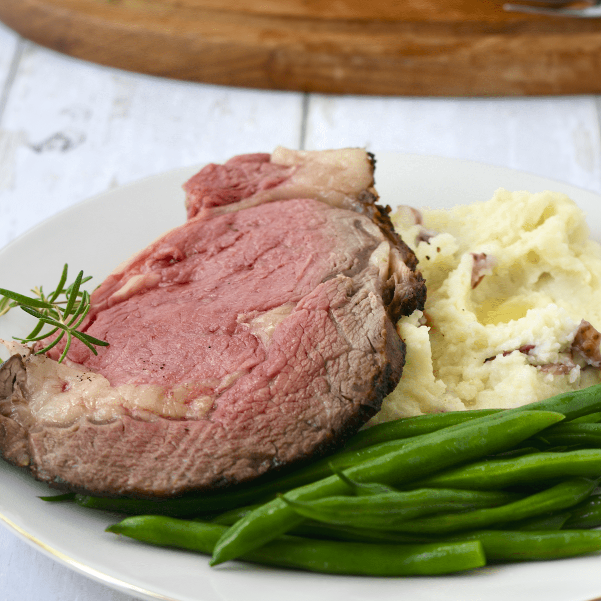 Slice of ribeye roast prime rib steak on white plate with green beans and mashed potatoes.