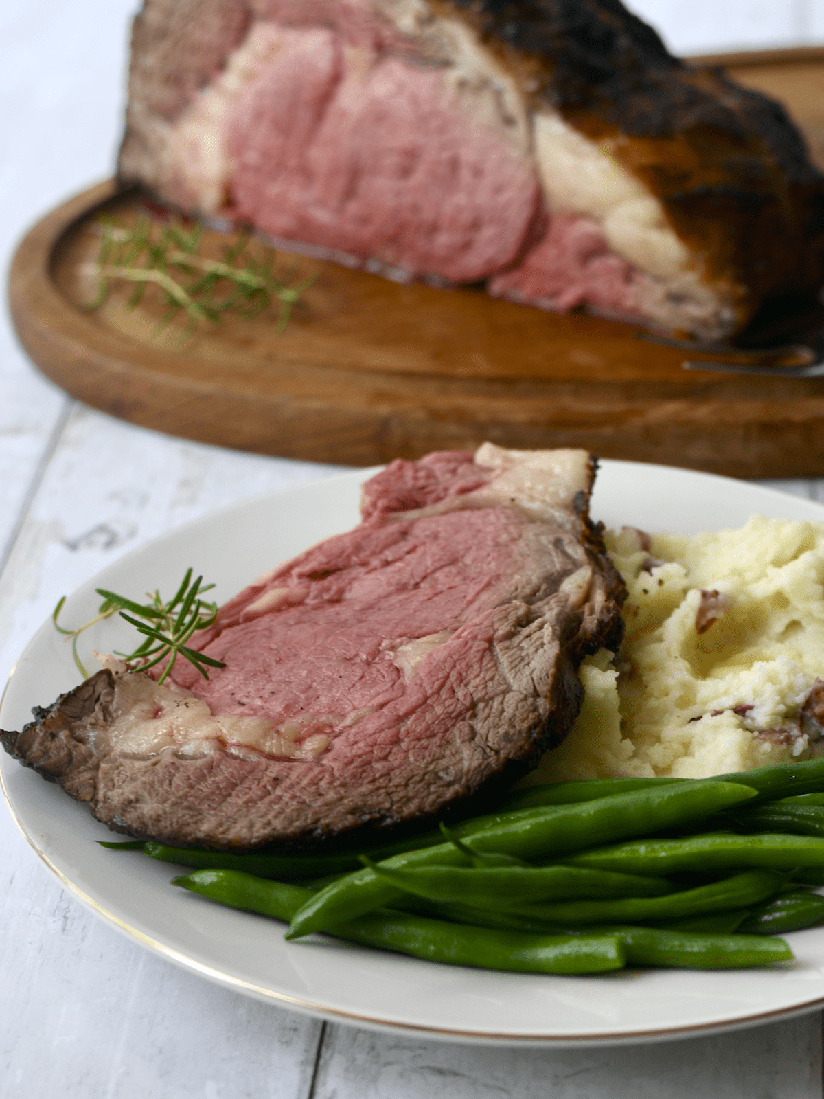 Boneless beef rib eye roast cooked in the background with a slice on a plate with green beans and mashed potatoes.