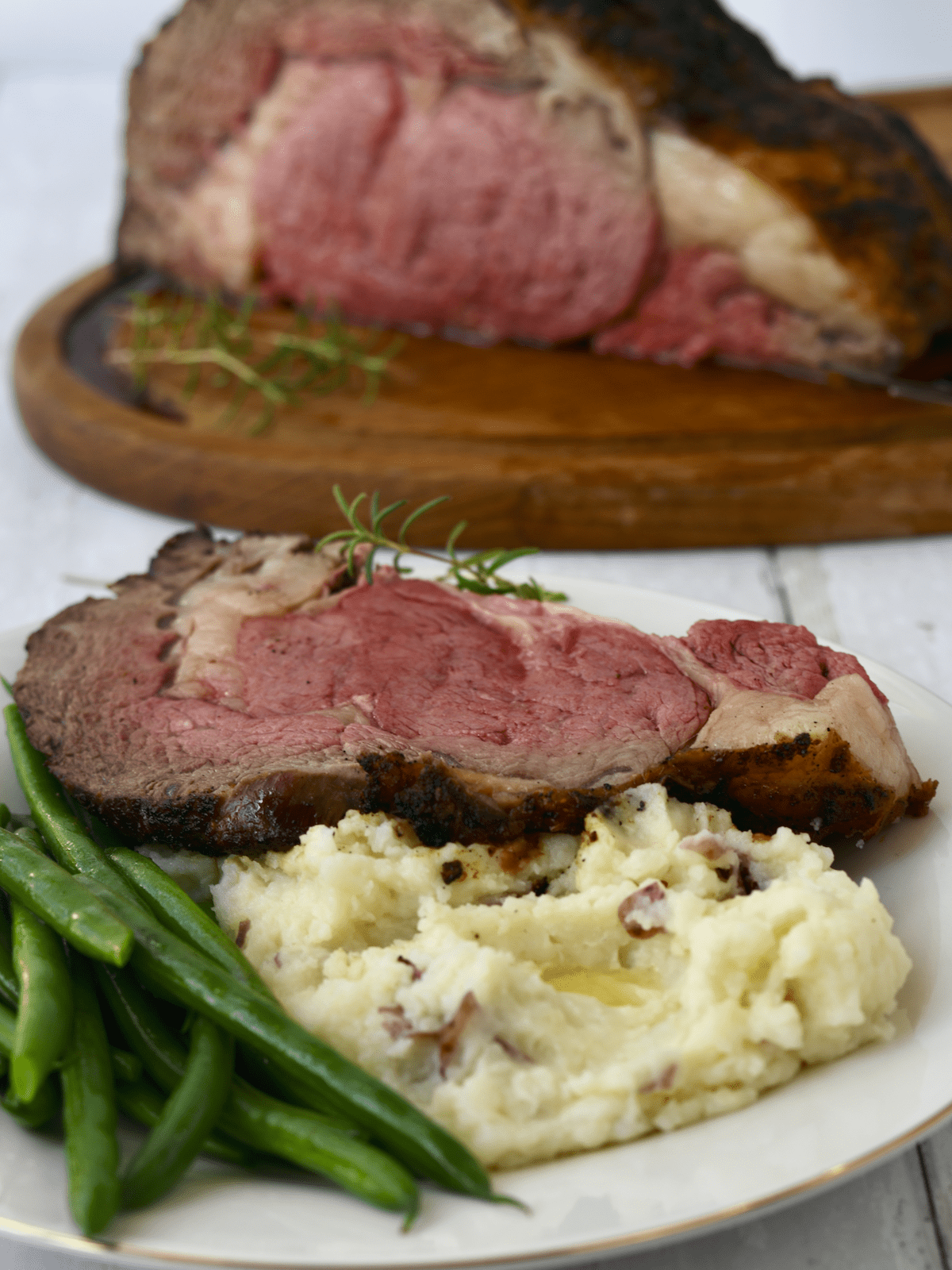 Standing ribeye roast in background with slice on plate with green beans and mashed potatoes.
