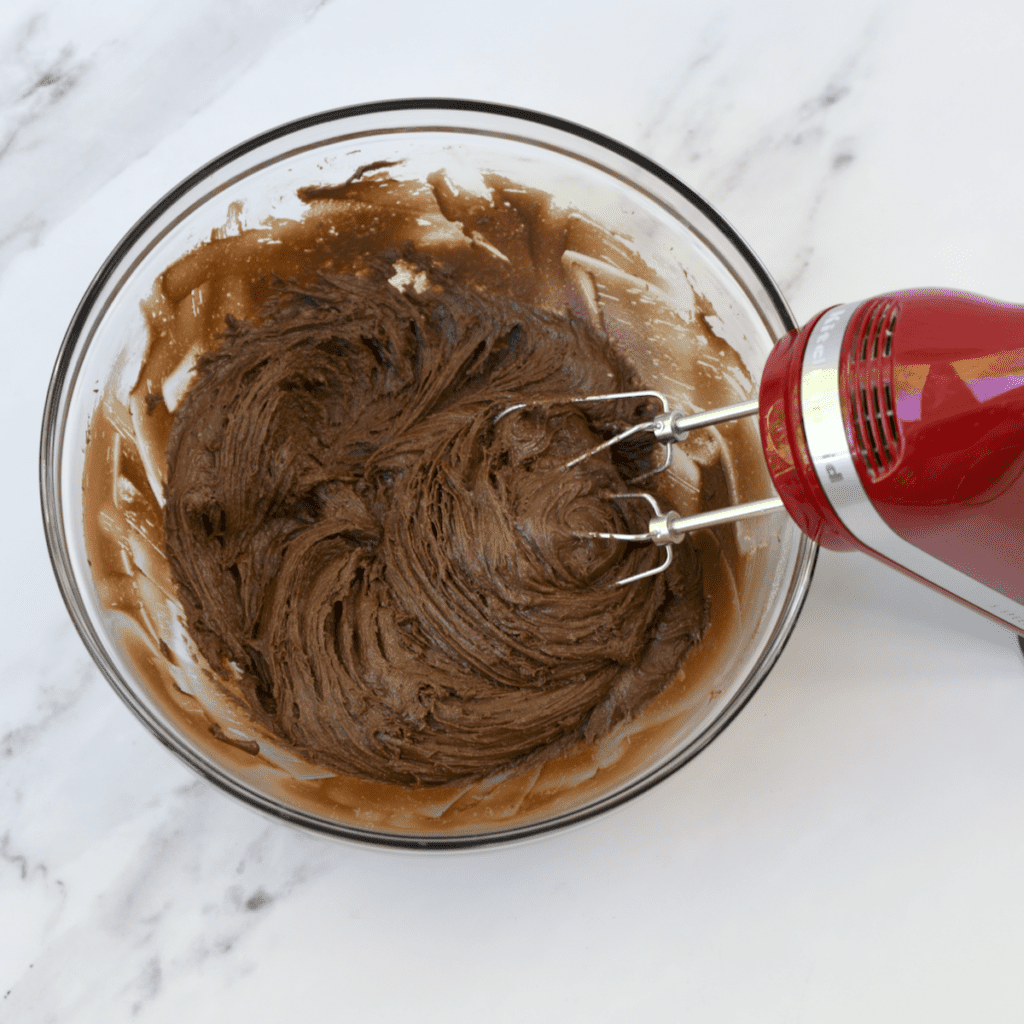 Glass bowl with  chocolate cake mix batter being mixed with red hand mixer.
