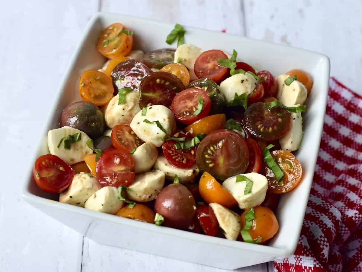 White bowl filled with dressed salad of cherry tomatoes, cubes of mozzarella cheese and shredded basil leaves.