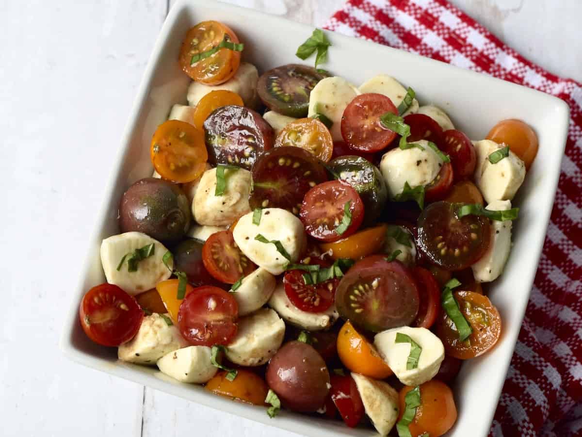 White bowl filled with dressed salad of cherry tomatoes, cubes of mozzarella cheese and shredded basil leaves.