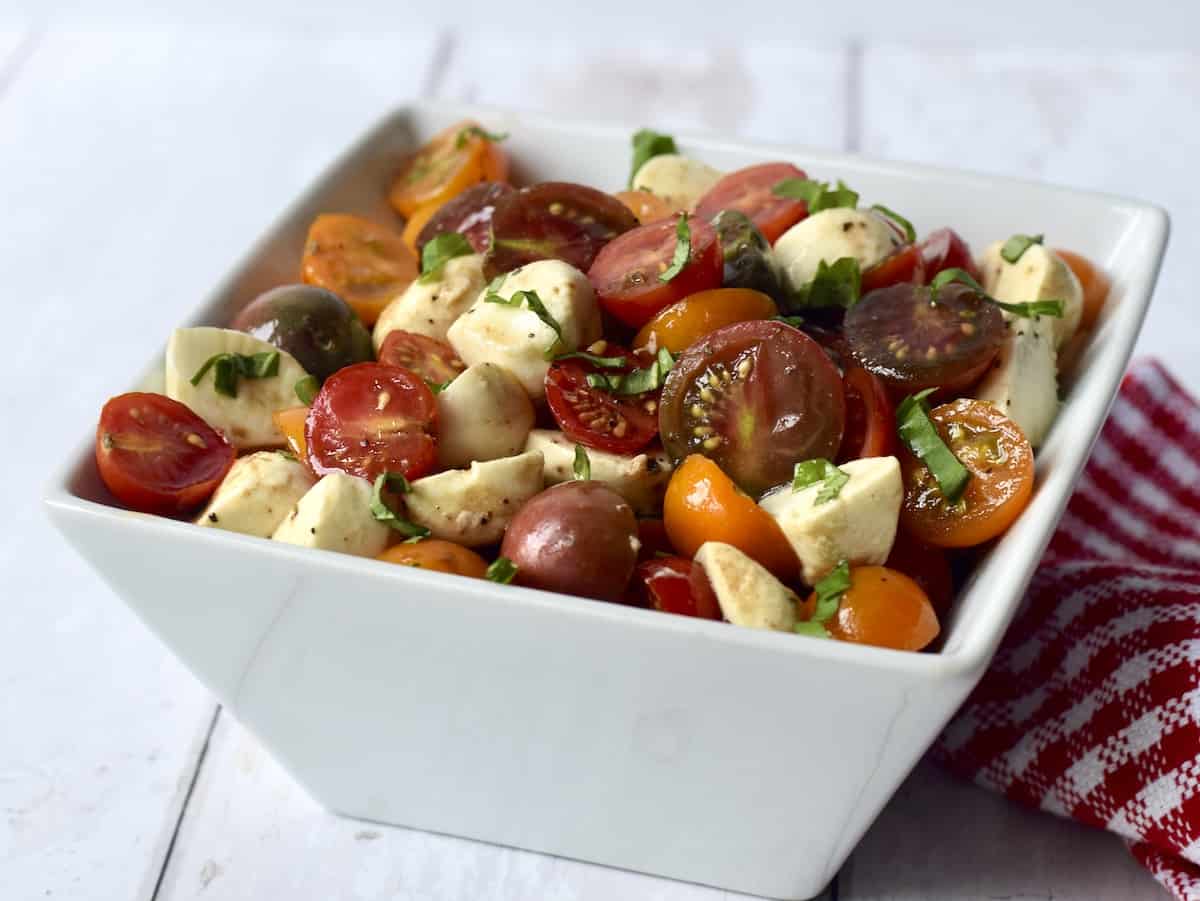 White bowl filled with dressed salad of cherry tomatoes, cubes of mozzarella cheese and shredded basil leaves.