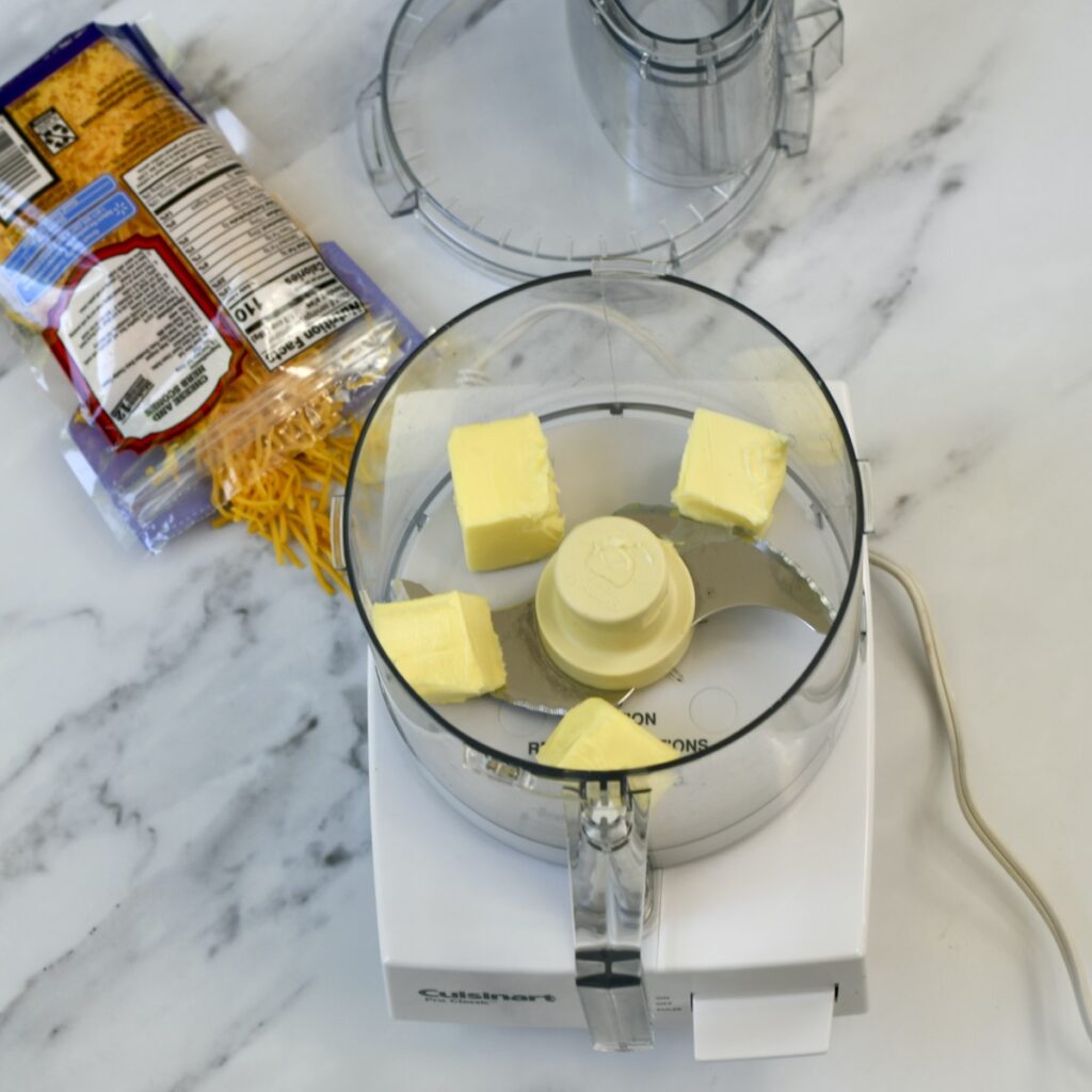 Butter cut in cubes in an open food processor with a bag of shredded cheese on surface next to it.

