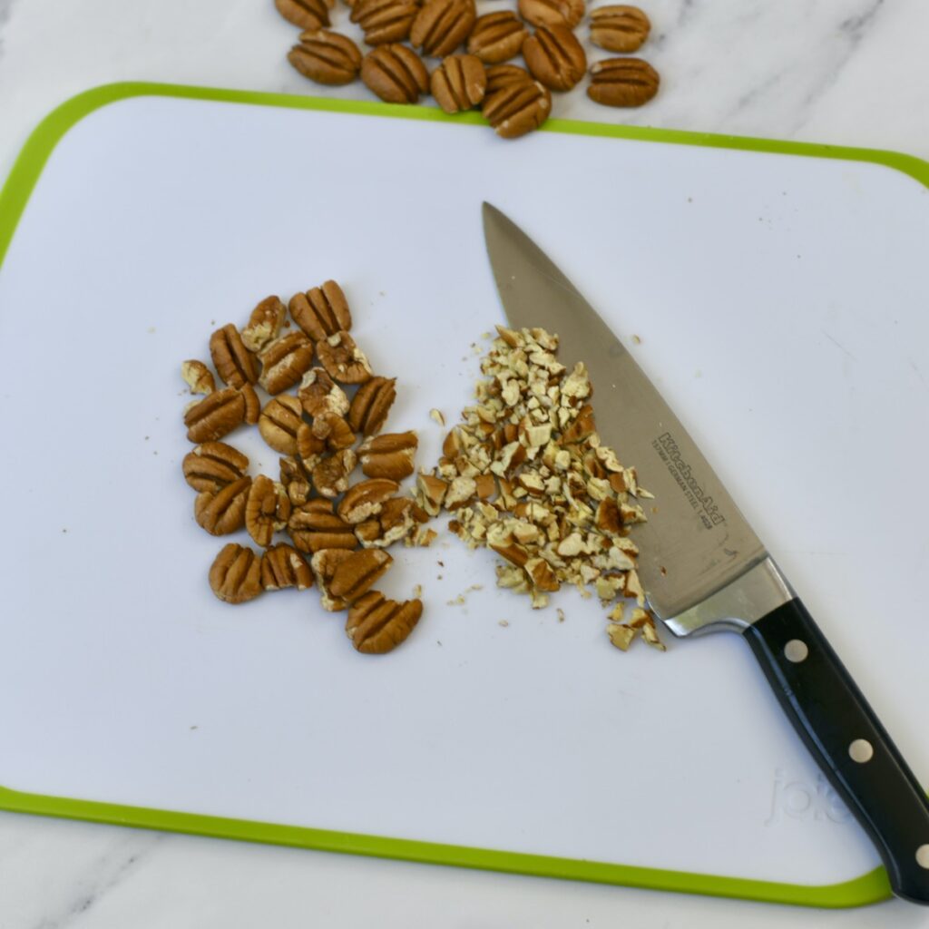 Chef knife next to chopped and whole pecans on a cutting board.
