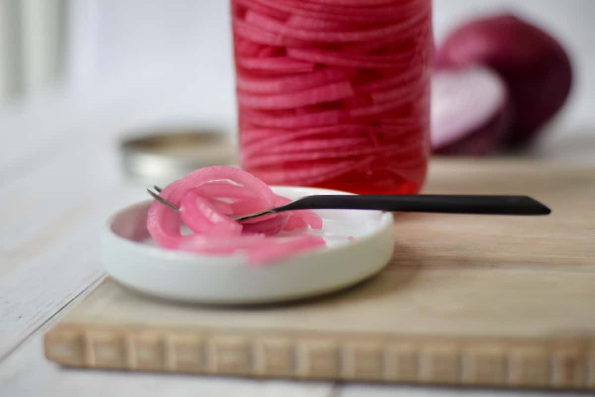 a fork with a portion pickled red onion set on a white plate with a jar of pickled onions in the background