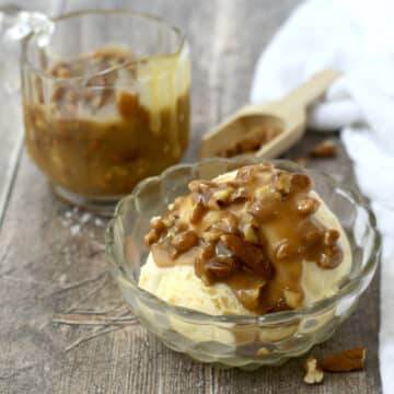 Pecan praline sauce drizzled on vanilla ice cream in a glass dish with glass sauce pitcher behind it.