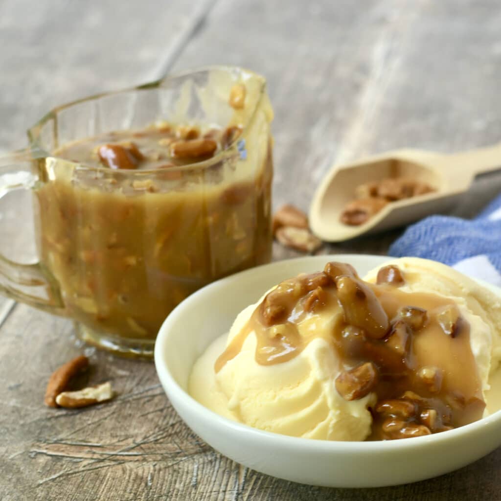 Praline Pecan Sauce drizzled over vanilla ice cream with glass pitcher of pecan caramel sauce behind it.