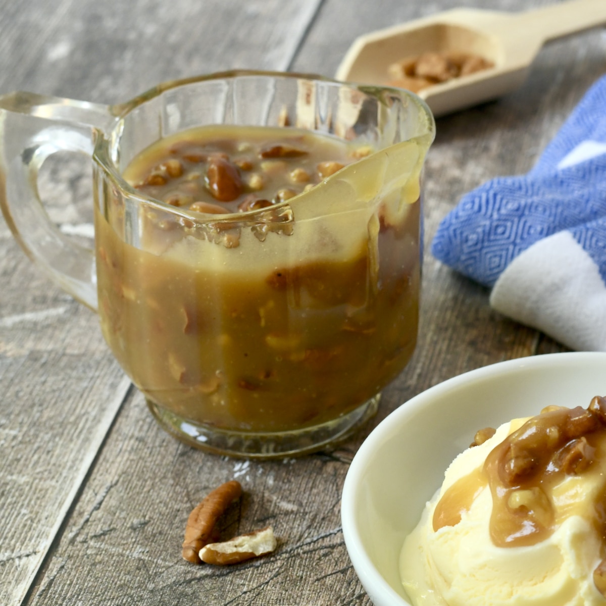 Glass syrup pitcher full of pecan praline sauce with drips and a bowl of ice cream in foreground.