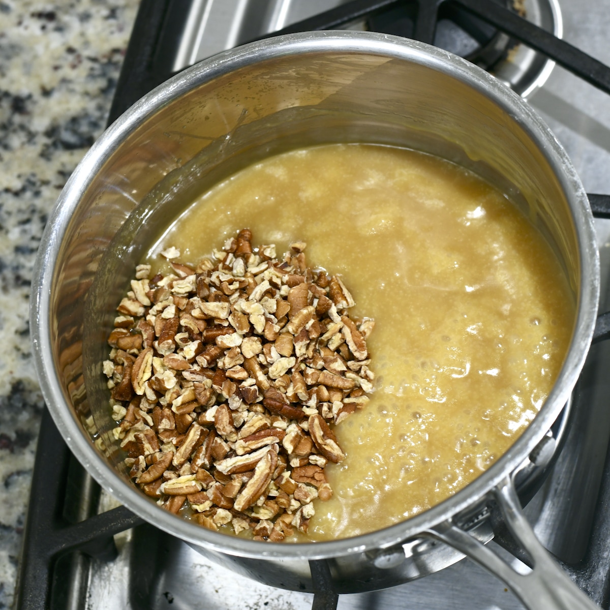 Praline sauce in a saucepan with chopped pecans added to the left side before stirring.