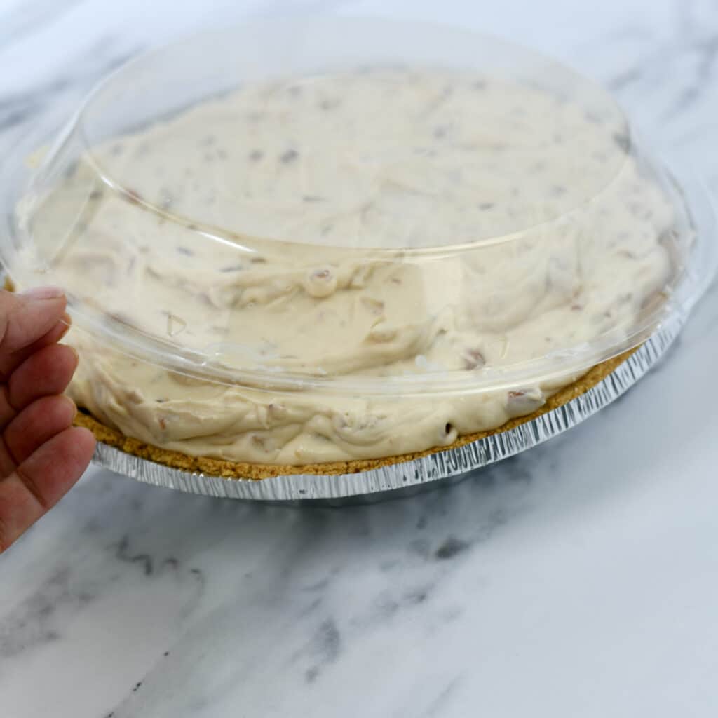 Hand placing the plastic lid from graham cracker crust on top of a pecan cream pie.
