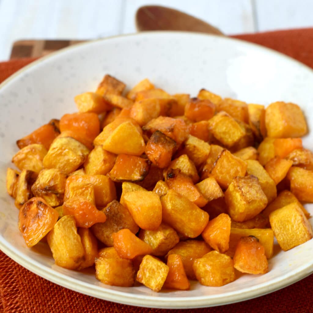 Bowl of roasted butternut squash on fall colored cloth with wooden spoon next to it.