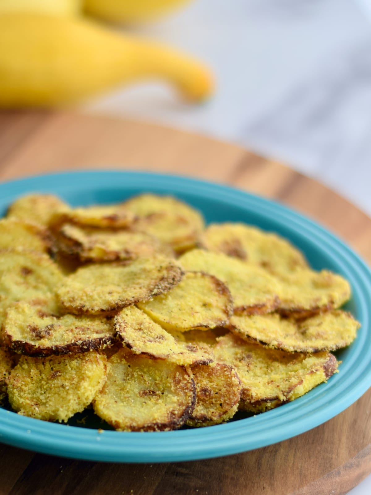 Blue plate of thinly sliced air fried yellow squash with squash in the background.