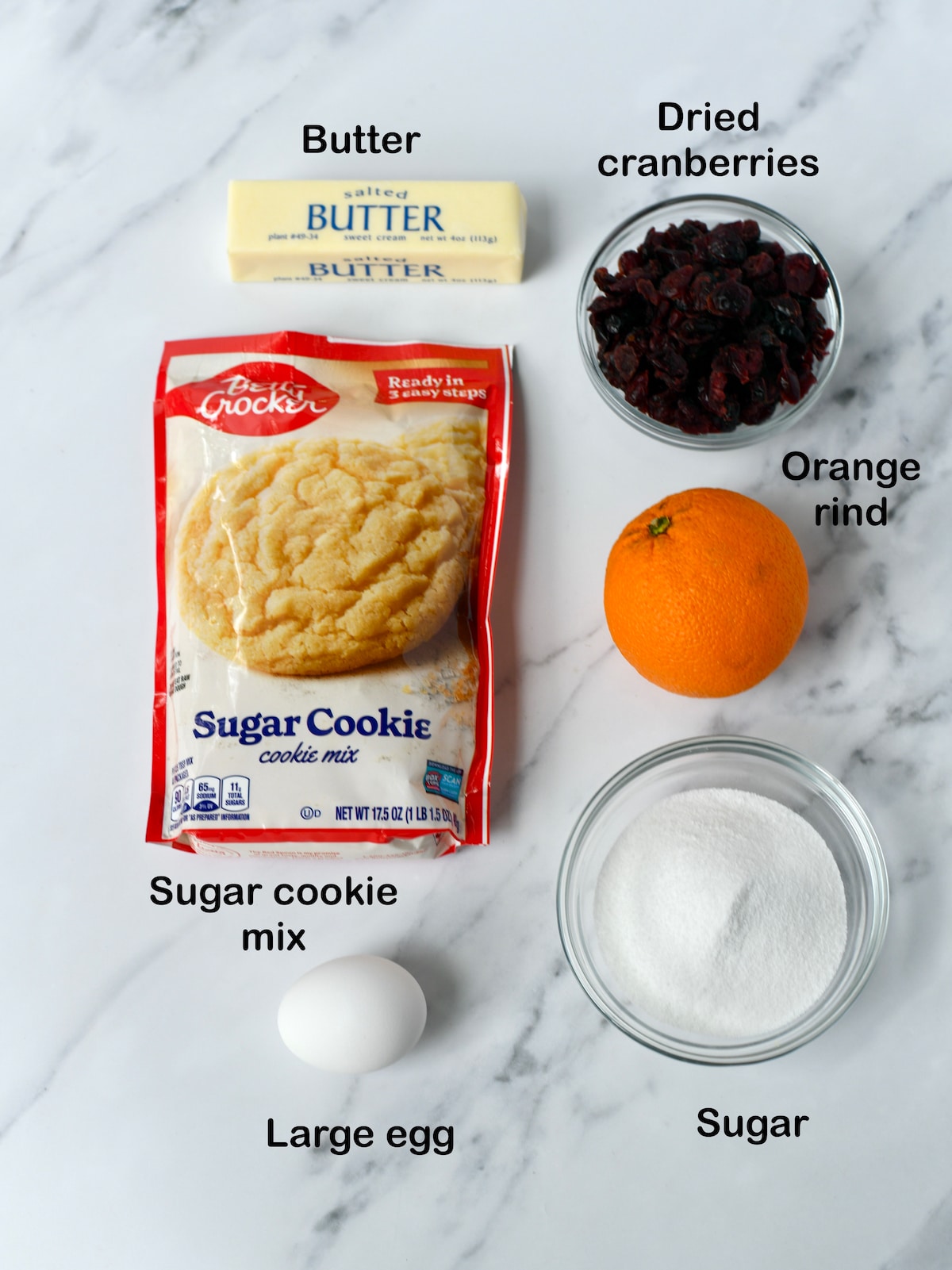 Labelled ingredients for cranberry orange cookies on a marble counter.